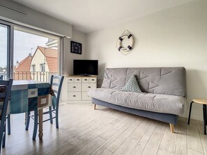 Un salon moderne avec un canapé gris, un téléviseur posé sur un meuble blanc, une table à manger avec des chaises, et une vue sur les bâtiments à l’extérieur.