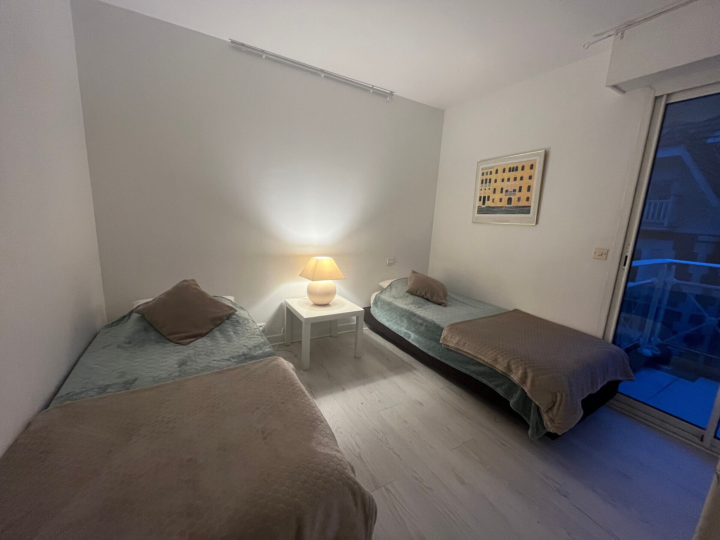 A small, well-lit room with two single beds, a bedside table with a lamp, and a framed picture on the wall.