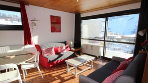 Een woonkamer met een rode bank, een grijze bank, een witte tafel en een witte stoel. Er is een raam met uitzicht op gebouwen en een balkon.