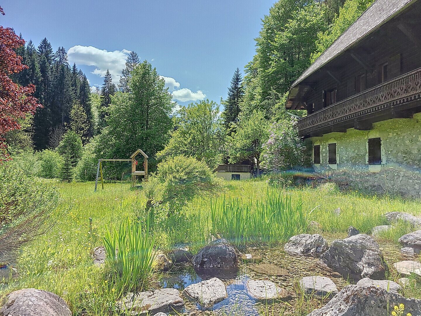 Un chalet rustique doté d’un balcon, entouré d’un jardin luxuriant et d’un petit étang.