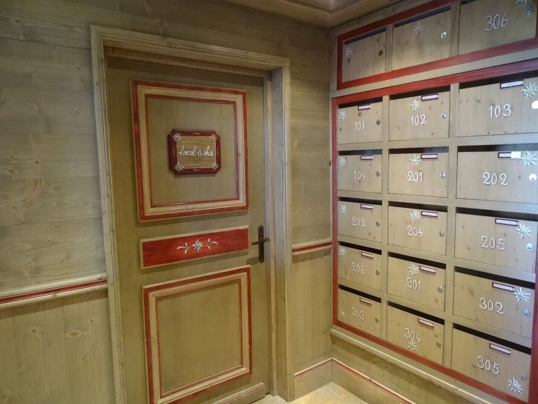 A row of wooden mailboxes with numbers, a door with a red frame, and a sign that reads 'local à ski.'