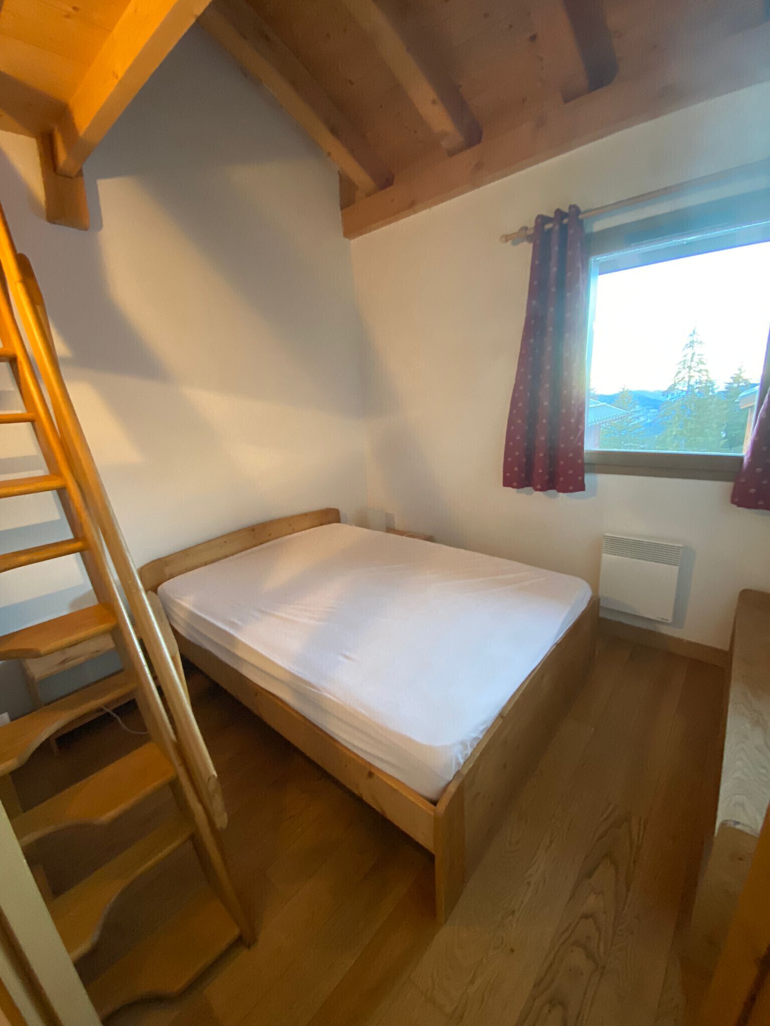 A small, single-bed room with a wooden ladder, a window with curtains, and a radiator.