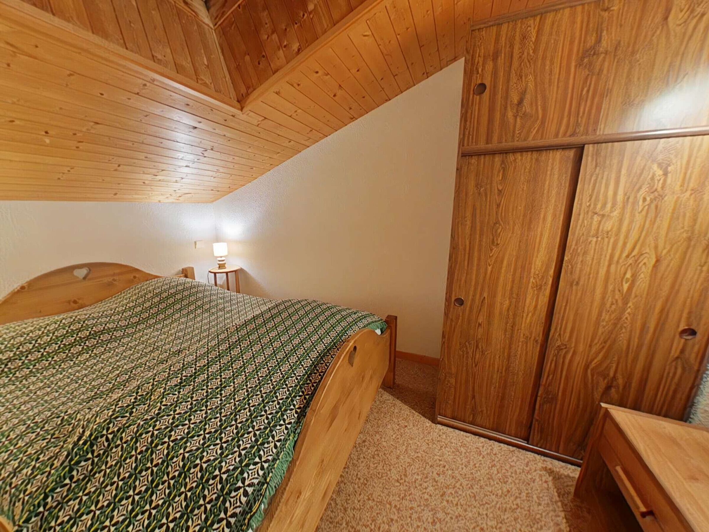 A bedroom with a wooden ceiling, a bed with a patterned bedspread, a nightstand with a lamp, and a wooden wardrobe.