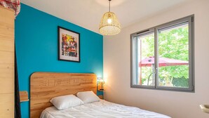 A bedroom with a wooden headboard, a bed with white bedding, a framed picture on the wall, a hanging light fixture, and a window with a view of greenery.