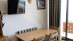 A wooden dining table with four chairs, a mounted television, a framed picture on the wall, and a window with curtains. - Cozy 1-Bedroom Apartment for 5 with South-Facing Balcony in L'Alpe d'Huez (Huez)