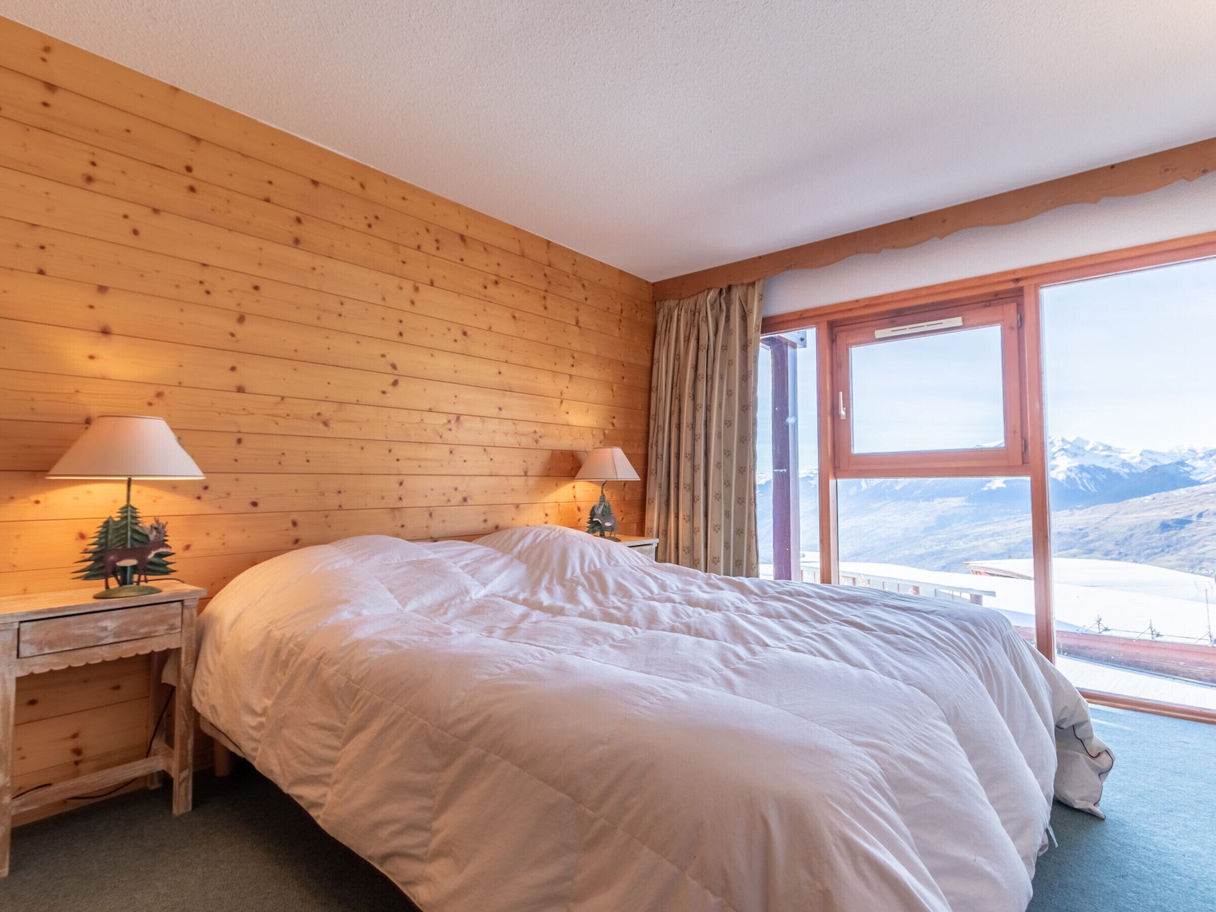 A bedroom with a large bed, wooden walls, two bedside lamps, a nightstand, and a window with a mountain view.