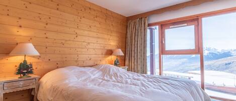 A bedroom with a large bed, wooden walls, two bedside lamps, a nightstand, and a window with a mountain view.