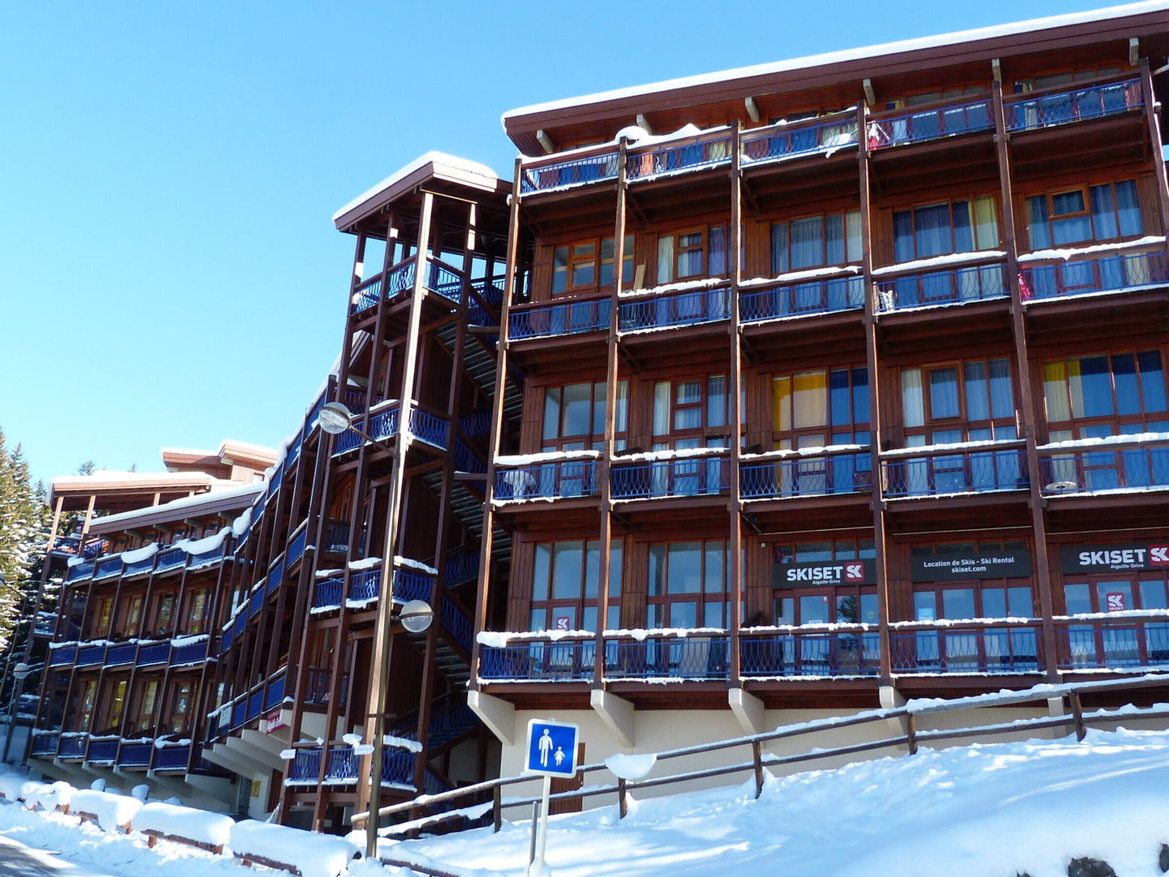 Un hôtel à plusieurs étages, doté de balcons et d’une enseigne indiquant un service de location de matériel de ski.
