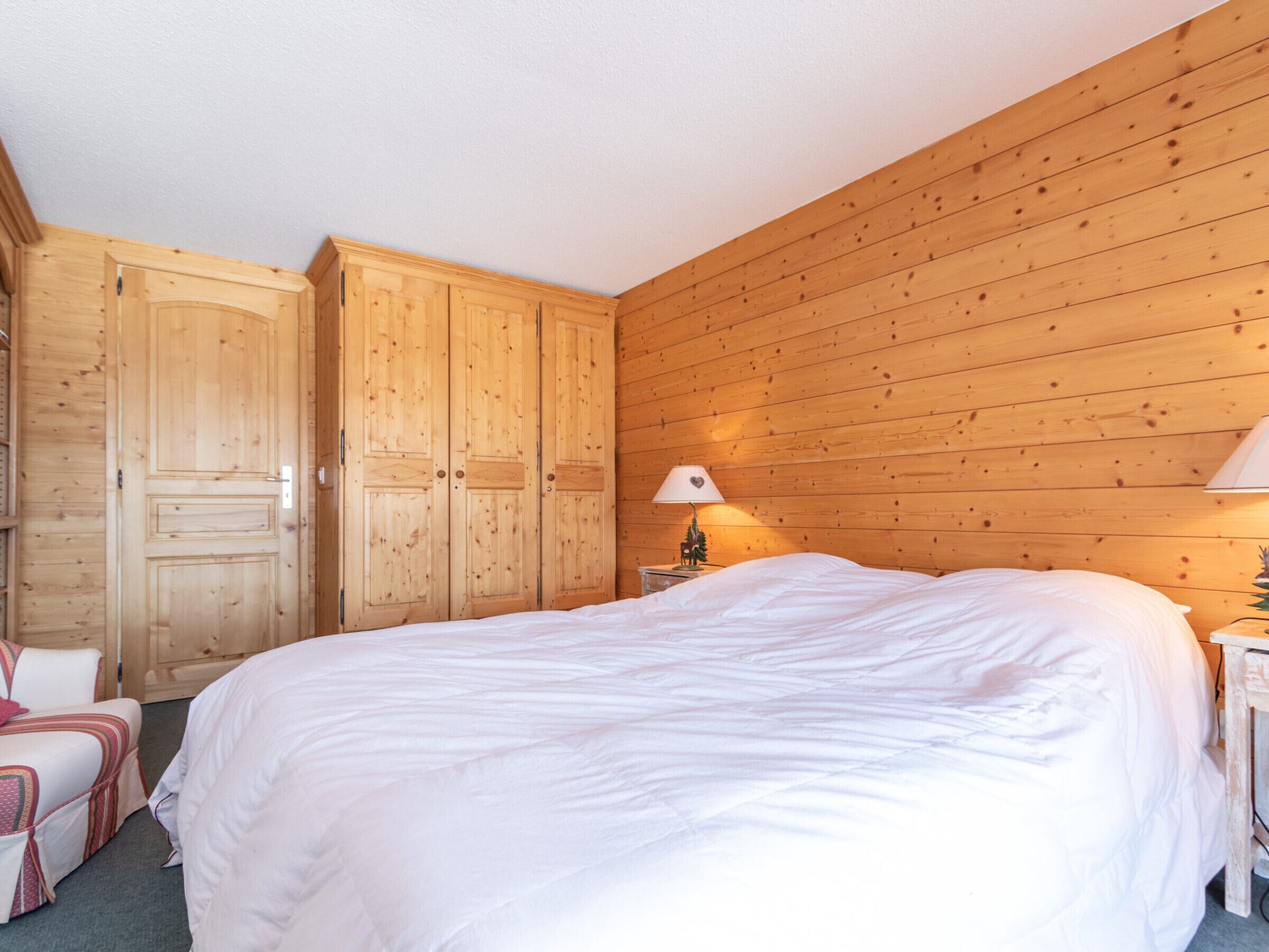 A bedroom with a large bed, wooden wardrobe, and a nightstand with a lamp.