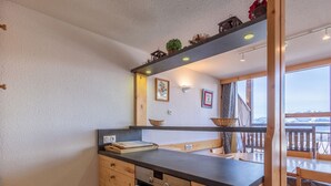 Une cuisine compacte avec des meubles en bois, un four encastré et un plan de travail agrémenté d’un panier et d’un tableau encadré.
