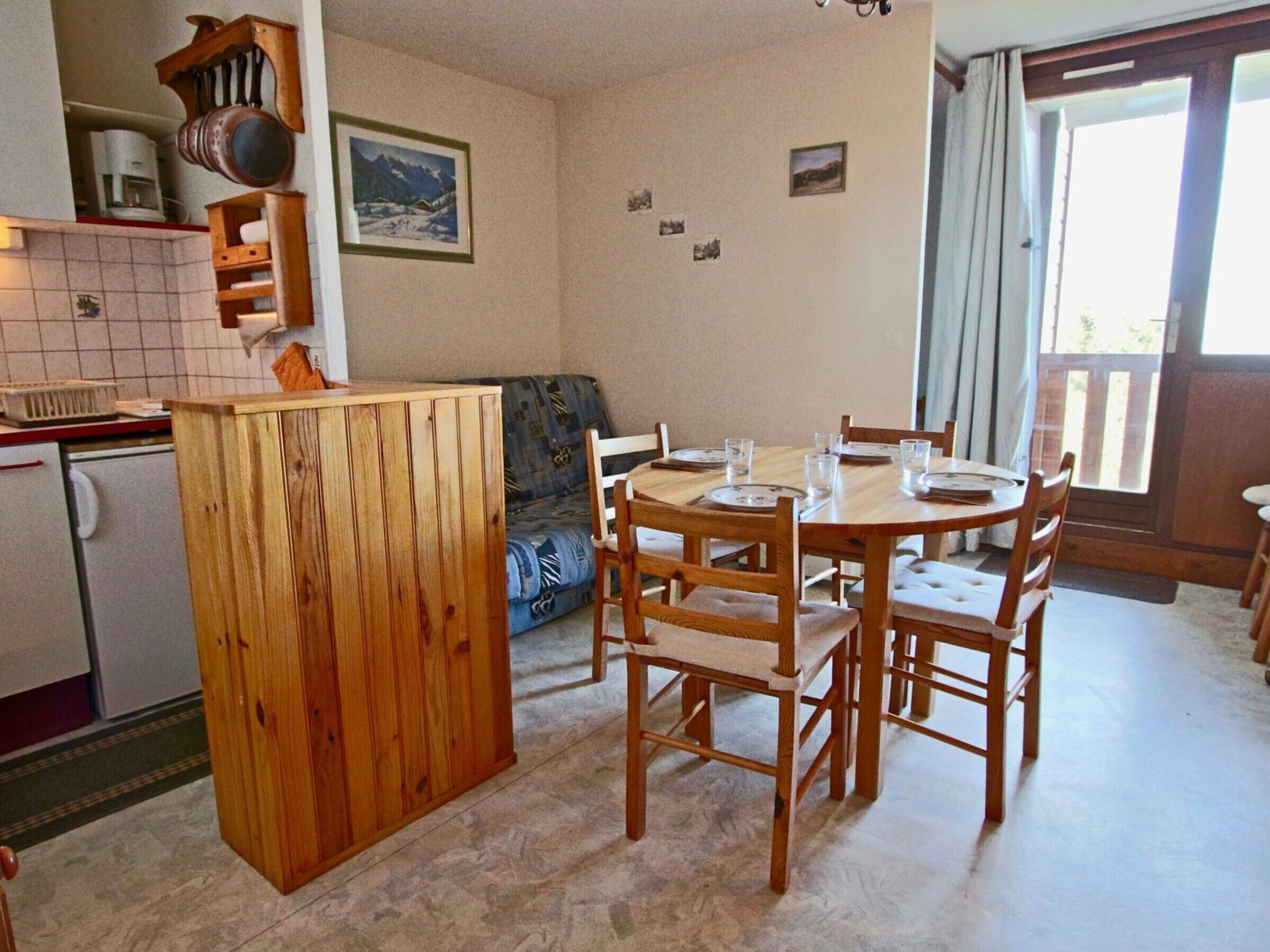 A compact kitchen with a wooden cabinet, a dining table with chairs, a sofa, and a refrigerator.