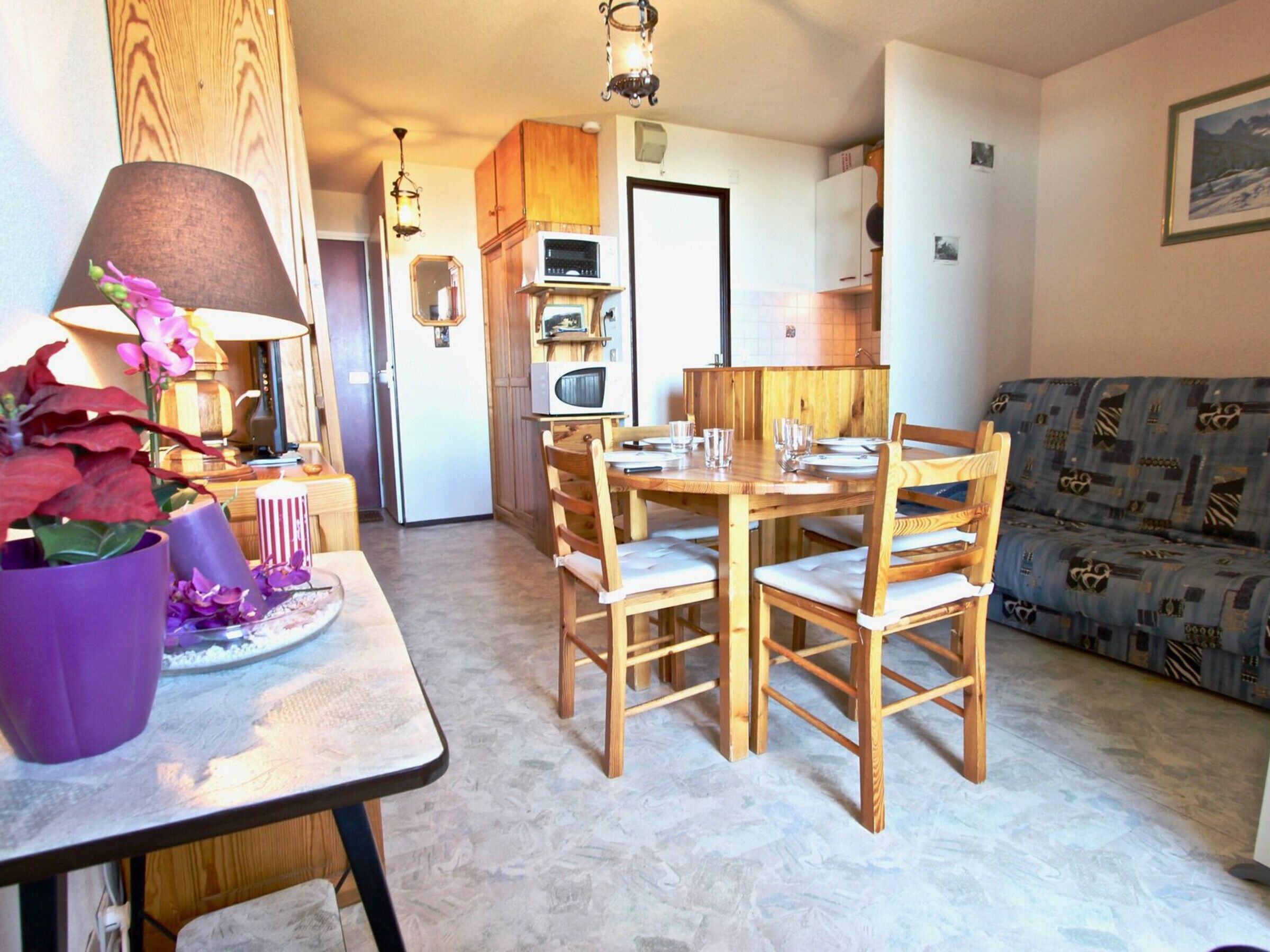 A cozy dining area with a wooden table and chairs, a small kitchen in the background, and a patterned sofa.