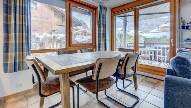 Un comedor con una mesa y sillas de madera, ventanas grandes con cortinas y vistas a un paisaje nevado de montañas.
