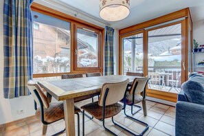 Uma sala de jantar com mesa e cadeiras de madeira, janelas amplas com cortinas e vista para uma paisagem montanhosa coberta de neve.