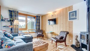 Una sala de estar acogedora con un sofá, un reposapiés y dos sillones, una televisión, una chimenea y una ventana con cortinas.