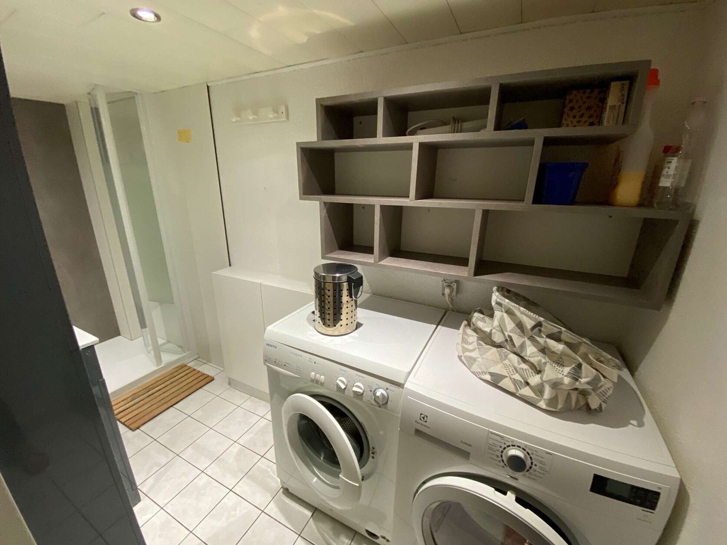 A compact laundry area with a front-loading washing machine, a drying machine, and a metal container on the countertop. Shelves above the machines are empty.