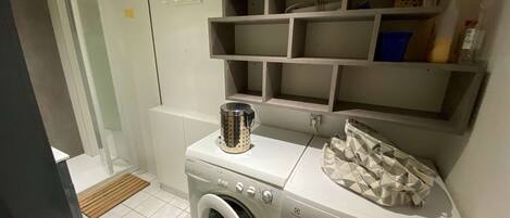 A compact laundry area with a front-loading washing machine, a drying machine, and a metal container on the countertop. Shelves above the machines are empty.