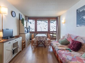 A cozy living area with a wooden floor, a sofa with a colorful blanket, a dining table with chairs, a TV on a cabinet, and a view of a snowy landscape through a window.