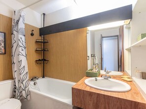 A bathroom with a wooden vanity, a bathtub, a shower curtain with a leaf pattern, a mirror, and a towel rack.