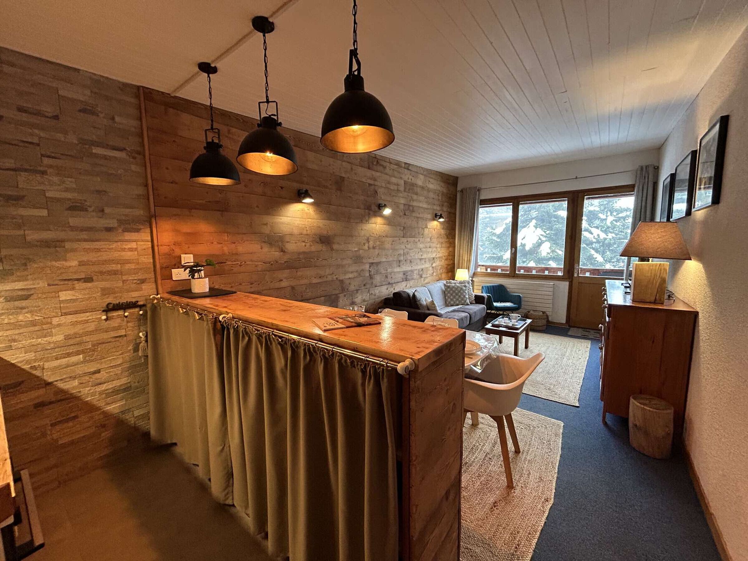 Eine gemütliche Küche mit einer Holztischinsel, einem Essbereich mit Sofa und Sesseln, und Blick auf eine verschneite Landschaft durch große Fenster.