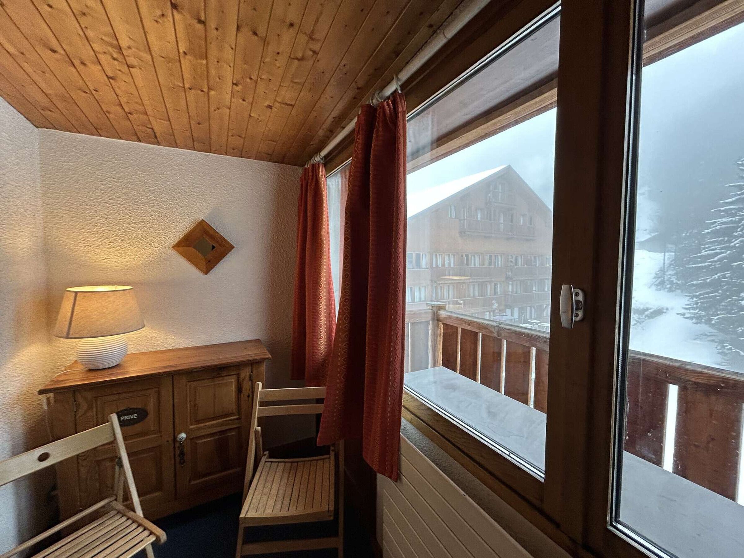 Une pièce chaleureuse avec un bureau en bois, une chaise, une lampe et une fenêtre ornée de rideaux rouges.