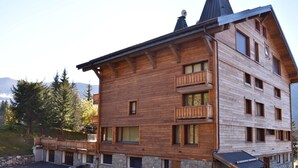 A multi-story wooden building with balconies, stone foundation, and a ski slope in the background.