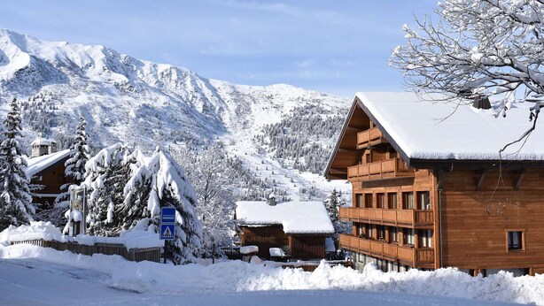 A wooden lodge with a balcony, surrounded by snow-covered trees and mountains.