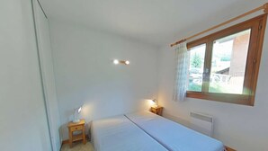 Une petite chambre propre, avec deux lits, une fenêtre avec des rideaux, une table de chevet avec une lampe et un radiateur.