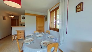 Un coin repas avec une table dressée pour six personnes, des chaises en bois, une lampe suspendue rouge, une fenêtre avec des rideaux et une porte en bois.