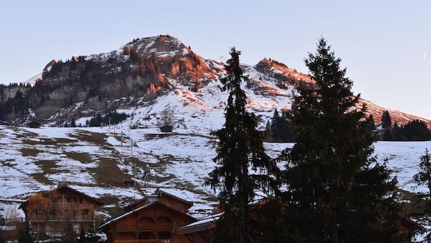 Un chalet en bois doté d’un balcon, entouré de montagnes et d’arbres couverts de neige.