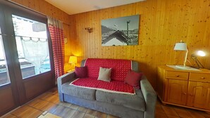 Ein gemütliches Zimmer mit einer holzverkleideten Wand, einem grauen Sofa mit roten und weißen Kissen und einem kleinen Holzschrank. Ein Fenster mit einem karierten Vorhang.