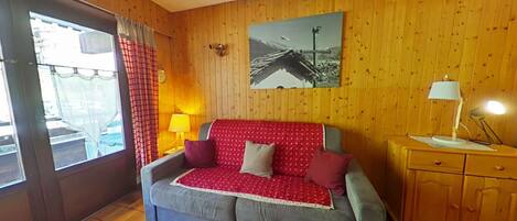 A cozy room with a wooden paneled wall, a grey sofa with red and white patterned cushions, a small wooden cabinet, and a window with a checkered curtain.
