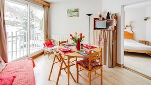 A cozy room with a bed, a dining table set for two, a TV, and a window with a view of trees. - Bright 2-room apt, sleeps 6, parking, near shops and skiing (Barèges)