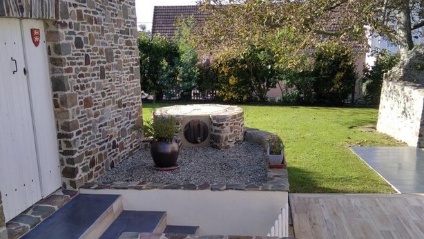 A stone wall with a potted plant, a small garden area with a circular stone feature, and a paved pathway leading to a building entrance.