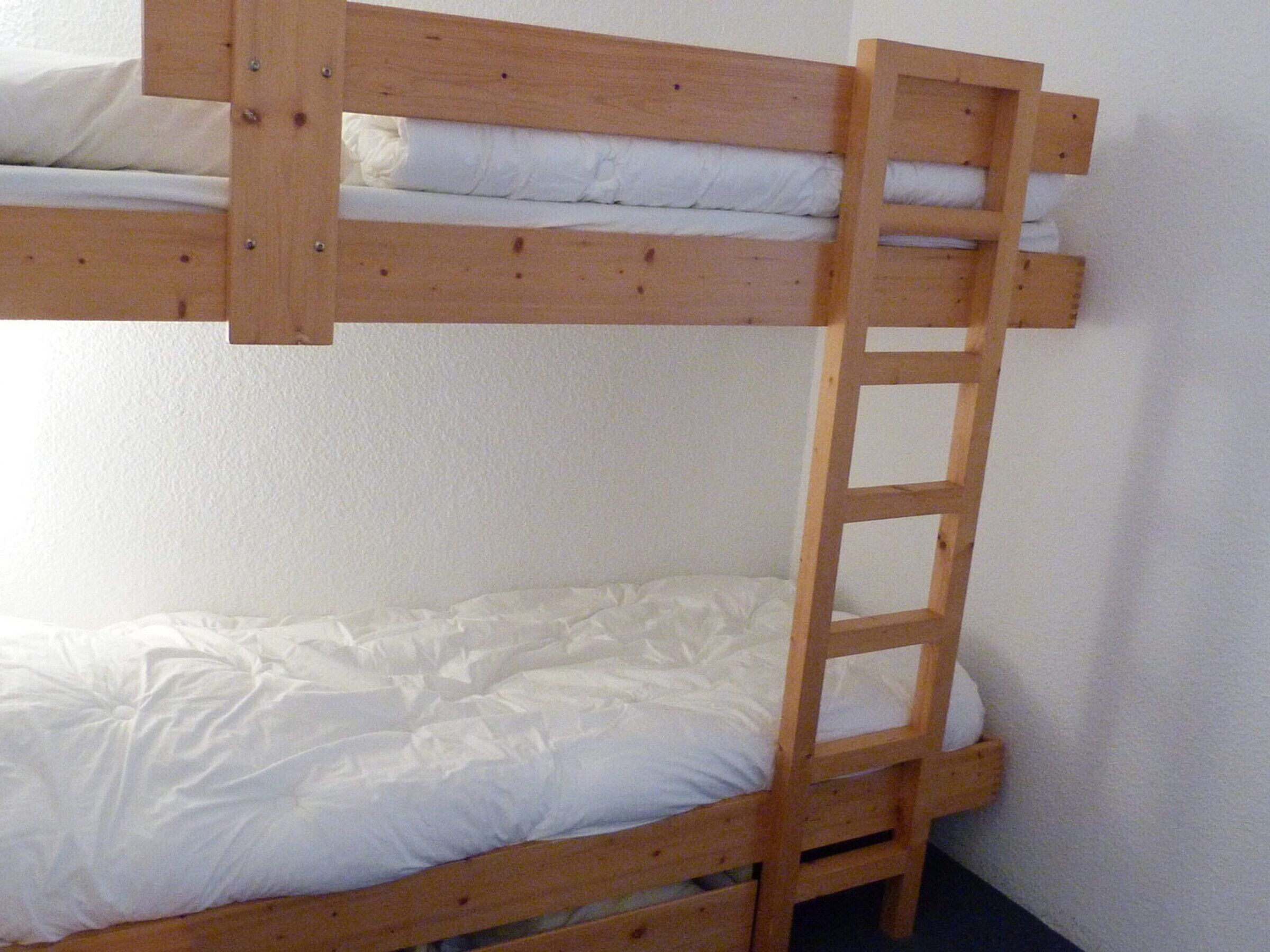 A bunk bed with a wooden ladder, white bedding, and a plain white wall.