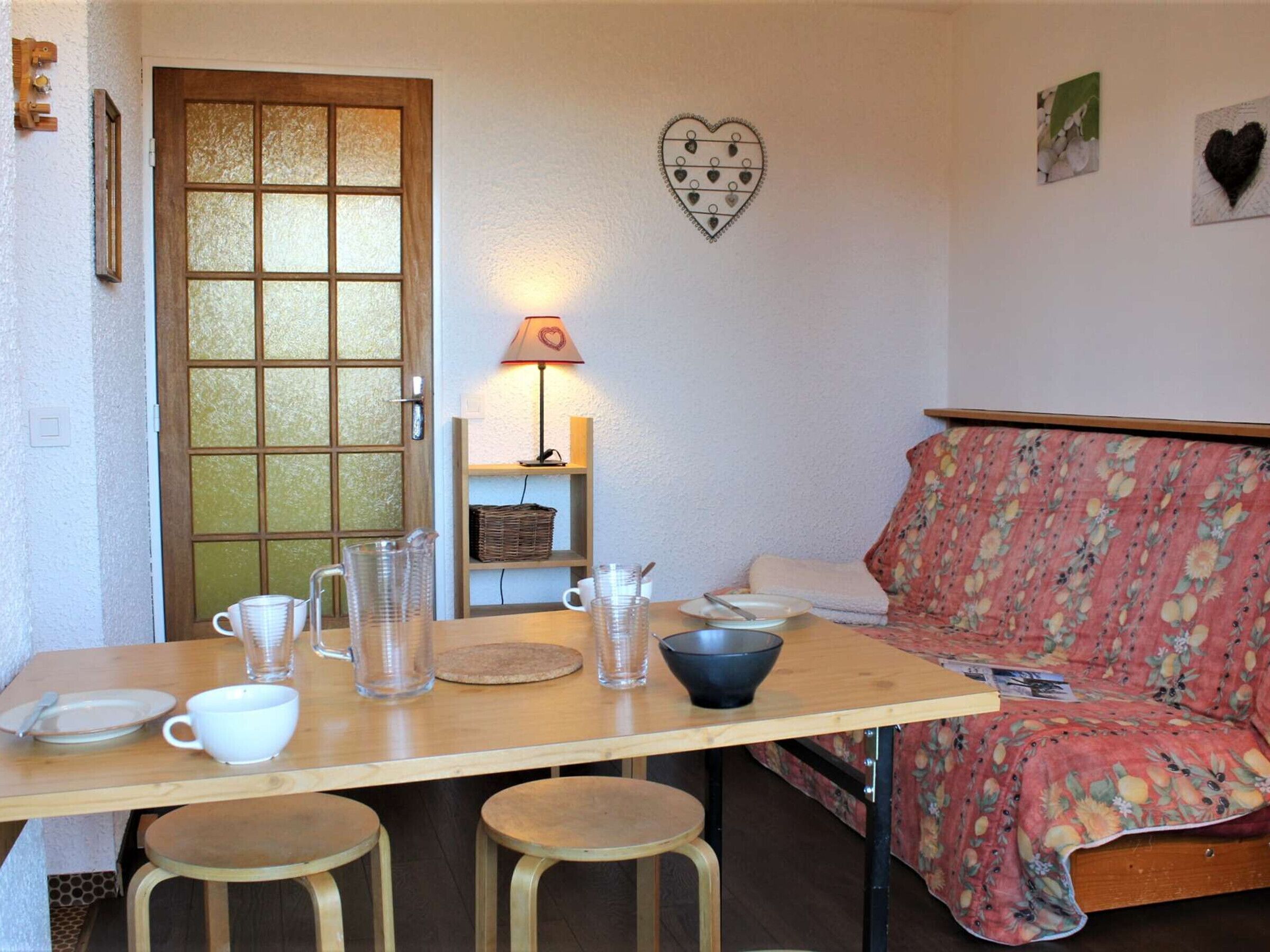 A dining area with a wooden table set for four, a floral sofa, and a wall-mounted lamp.