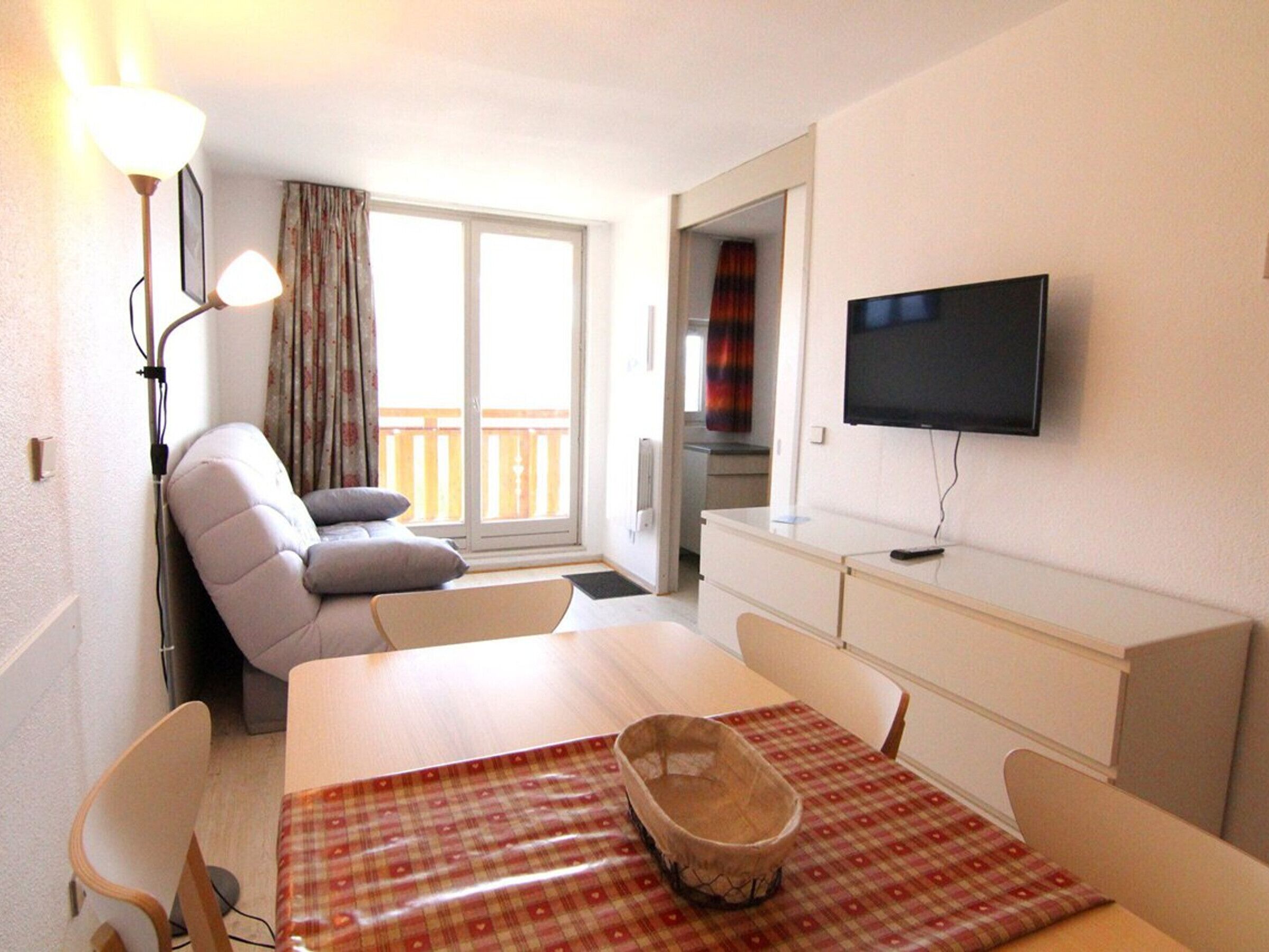Habitación de hotel con sofá, una mesa de comedor con mantel a cuadros, televisor montado en la pared y vistas al balcón exterior.