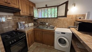 Une cuisine compacte équipée d’une cuisinière, d’un évier et d’un lave-linge, de meubles en bois et d’une fenêtre donnant sur un espace verdoyant.