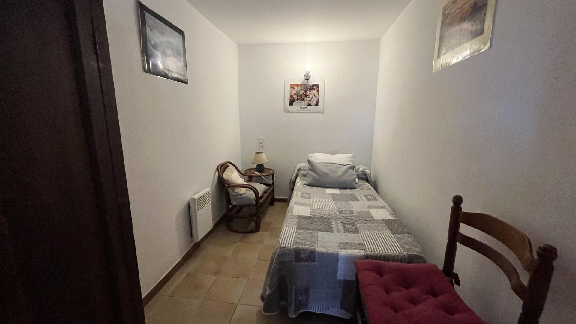 Ein kleines, schmales Zimmer mit einem Bett, einem Sessel, einem Nachttisch und einer Wandlampe.