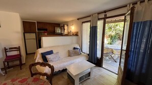 Un salon confortable avec un canapé, une petite table, un fauteuil, un coin cuisine et une porte-fenêtre coulissante donnant sur une terrasse extérieure.