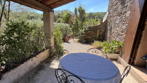 Une terrasse couverte avec une table ronde et des chaises, entourée de plantes en pot et d’un mur de pierre.