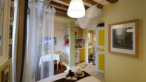Une pièce comprenant un lit, une table avec un appareil photo et un chapeau, un téléviseur, et un coin cuisine visible à travers une porte ouverte.