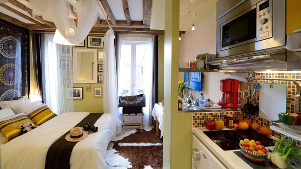 Une pièce comprenant un lit, une petite table avec un sac à main, une cuisine avec une cuisinière et un four à micro-ondes.