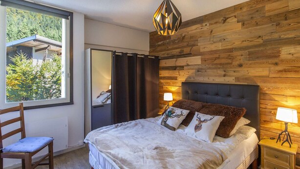 A bedroom with a large bed, a wooden wall, a window, and a mirror. - Spacious 3-Bedroom Apartment for 8 – Near Ski Slopes in Gourette, Fully Equipped (Gourette)