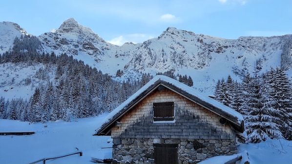 A stone cabin with a wooden roof, surrounded by snow-covered trees and mountains.