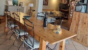 A dining area with a wooden table and chairs, a kitchen in the background, and a high ceiling with exposed beams. - Camfier self-catering cottage (TOURVES)