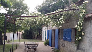 Zona exterior rústica amb taula i cadires, una pérgola coberta de flors blanques i un edifici amb persianes blaves.