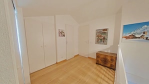 A spacious room with wooden flooring, white walls, and a wooden chest. There are white wardrobes and a framed picture on the wall.