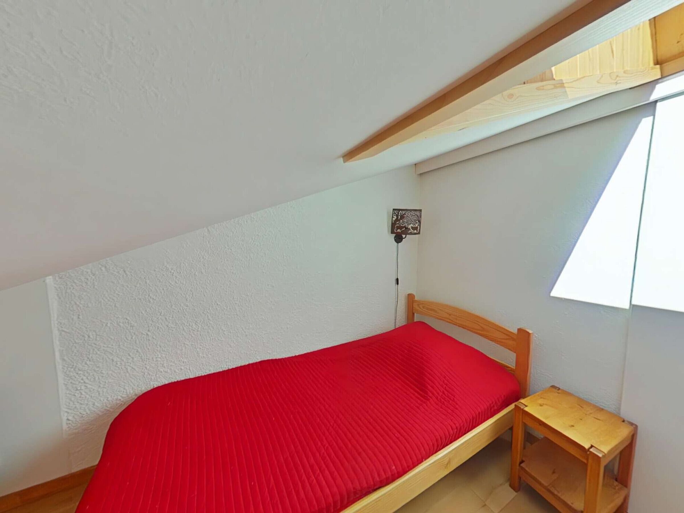 A small, single-bed room with a sloped ceiling and a red bedspread.
