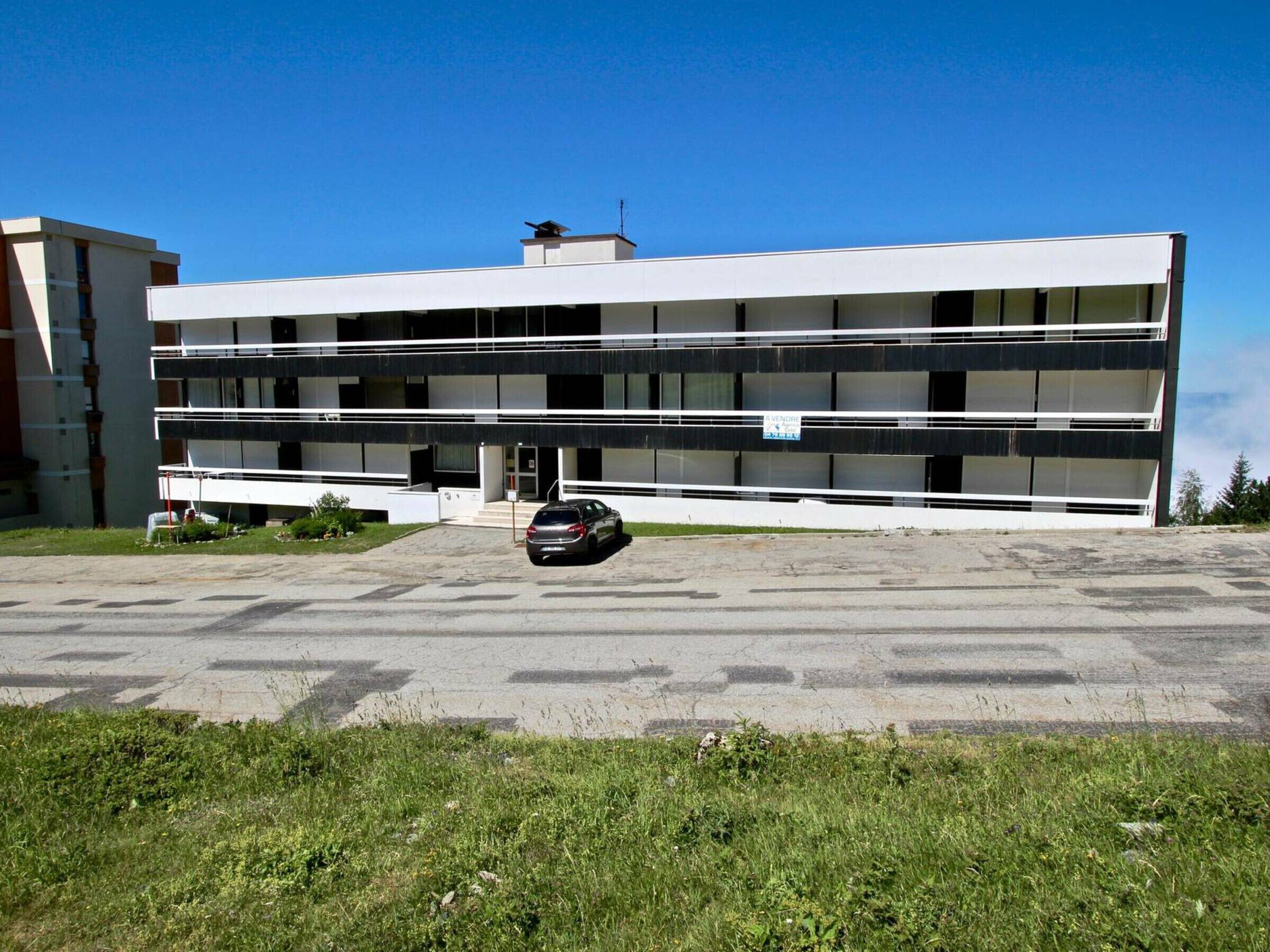 Monikerroksinen rakennus, jossa on tasakatto, sen edessä on pysäköity auto ja sen pohjakerroksessa on kyltti.